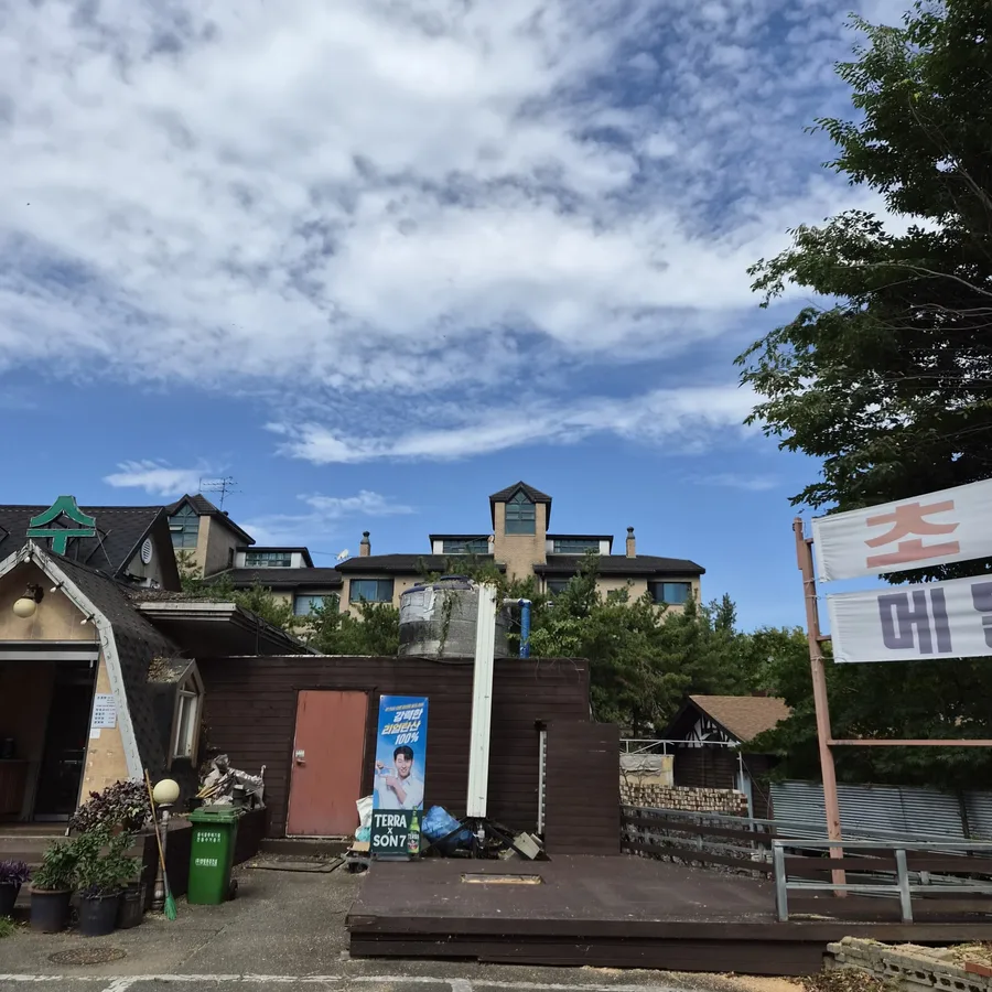 건물과 주차장이 보이는 넓은 전경 사진. 오른쪽에 잎이 적은 나무와 현수막 일부가 보이나 작업 장면은 없음.